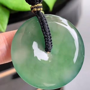 Pu&ograve; includere: Un ciondolo di giada verde su una collana di cordoncino nero. Il ciondolo &egrave; rotondo e ha un foro al centro.
