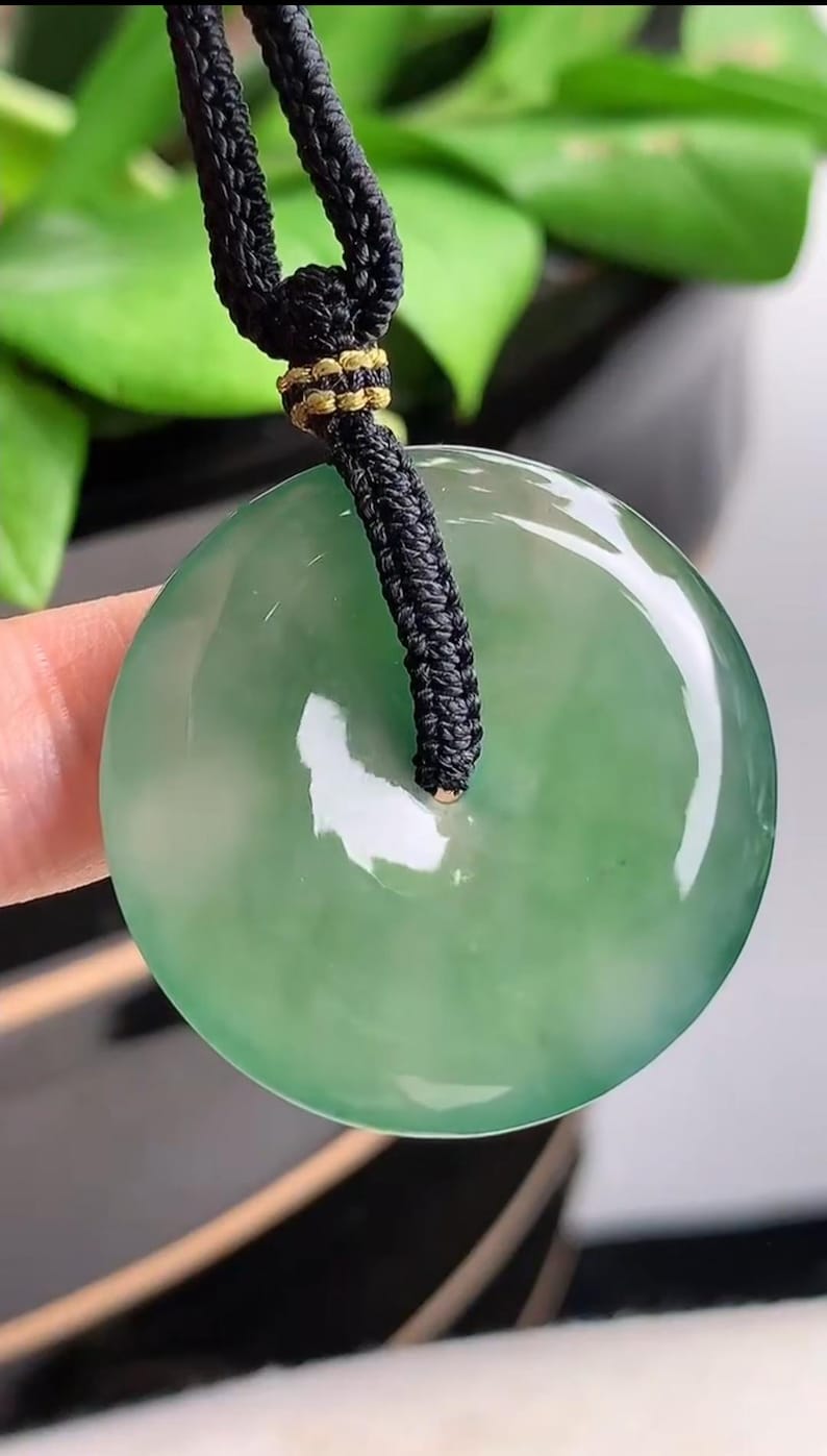 Pu&ograve; includere: Un ciondolo di giada verde su una collana di cordoncino nero. Il ciondolo &egrave; rotondo e ha un foro al centro.
