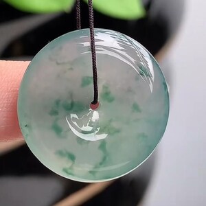 Puede incluir: Colgante redondo de jade transl&uacute;cido con un dise&ntilde;o veteado verde y blanco. Cuelga de un cord&oacute;n marr&oacute;n oscuro. El jade tiene un acabado pulido y brillante. El fondo est&aacute; borroso, con follaje verde visible.