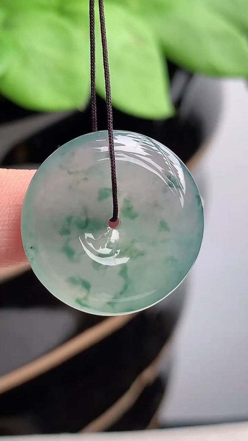 Puede incluir: Colgante redondo de jade transl&uacute;cido con un dise&ntilde;o veteado verde y blanco. Cuelga de un cord&oacute;n marr&oacute;n oscuro. El jade tiene un acabado pulido y brillante. El fondo est&aacute; borroso, con follaje verde visible.