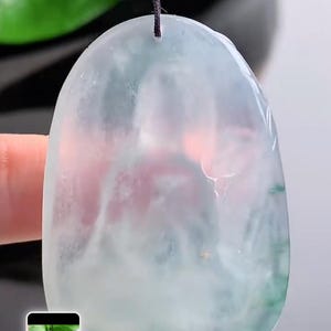 May include: A jade pendant with a rounded shape, suspended from a black cord. The jade features a blend of pale green, white, and hints of pink. The pendant is smooth and polished, reflecting light.
