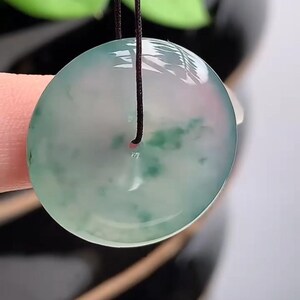 Puede incluir: Colgante redondo de jade transl&uacute;cido con un cord&oacute;n oscuro. El colgante tiene un tono verde claro y rosa con inclusiones verdes m&aacute;s oscuras. El jade es liso y pulido, reflejando la luz. El fondo est&aacute; borroso.