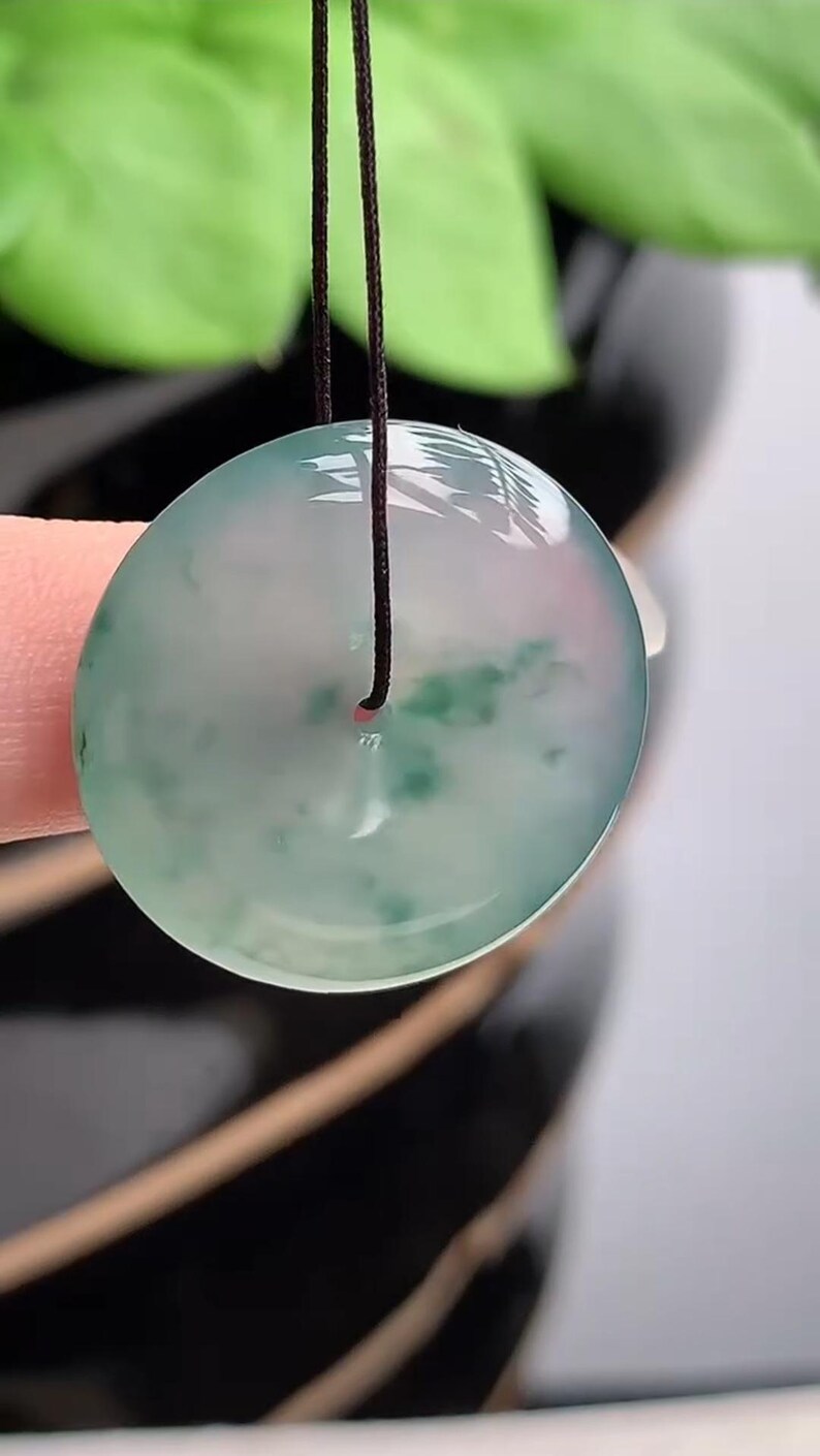 Puede incluir: Colgante redondo de jade transl&uacute;cido con un cord&oacute;n oscuro. El colgante tiene un tono verde claro y rosa con inclusiones verdes m&aacute;s oscuras. El jade es liso y pulido, reflejando la luz. El fondo est&aacute; borroso.
