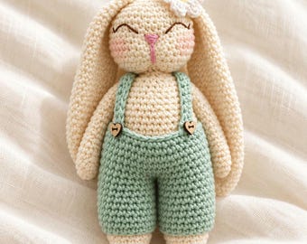 Könnte beinhalten: Eine cremefarbene Häkel-Hasenpuppe mit langen Ohren, die eine salbeigrüne Latzhose mit zwei kleinen herzförmigen Holzknöpfen trägt. Der Hase hat gestickte geschlossene Augen, eine rosa Nase, rosige Wangen und eine weiße Gänseblümchenblüte am Kopf.