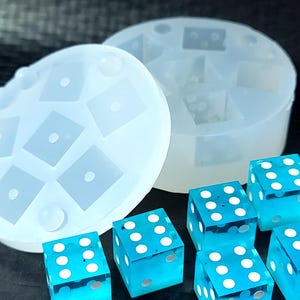 May include: A translucent silicone mold for casting dice, featuring a circular lid with square indentations and a cylindrical base, accompanied by four transparent blue six-sided dice with white pips on each face, arranged in a cluster.