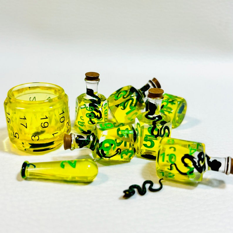May include: A set of yellow glass dice with green numbers and snake designs. The dice are in the shape of bottles and are arranged on a white surface. One die is a 20-sided die with black numbers.