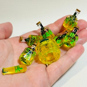 May include: A set of seven yellow dice with black numbers and green accents. The dice are shaped like small bottles and are arranged in a hand.