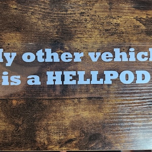 May include: A white sticker on a brown wooden surface with the text "My other vehicle is a HELLPOD".