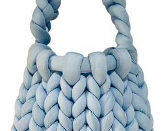 May include: A pale blue, hand-knitted handbag with a braided handle. The bag has a chunky knit design and a small gold-coloured metal tag with the initials "E J". The bag is a soft, pastel blue colour.