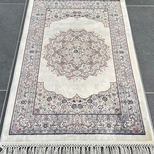 May include: A white prayer rug with a detailed floral pattern in shades of pink, brown, and gray. The rug has a fringe border.