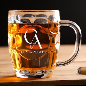 May include: A clear glass beer mug filled with amber liquid, featuring the initials "CA" and the name "Clark Austin". The mug sits on a wooden surface, with a lit match and a cigar nearby. The background is dark.