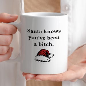 May include: White ceramic mug with the text "Santa knows you've been a bitch." The mug features a red and white Santa hat graphic. The mug is held by a person.