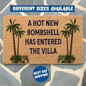 May include: A rectangular tan coir doormat featuring two green palm tree illustrations on the left and right sides. The text in black reads A HOT NEW BOMBSHELL HAS ENTERED THE VILLA. The mat has a coarse, natural fiber texture and a clean, flat design.