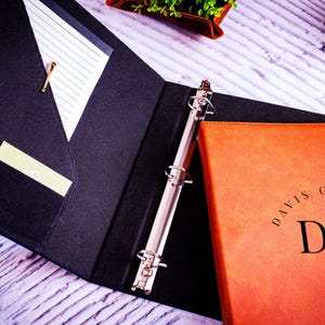 May include: A black binder with a white lined notepad and a silver binder mechanism. The binder is open to reveal a brown leather folder with the text "DAVIS GARGOYLE D/G" embossed on the front.