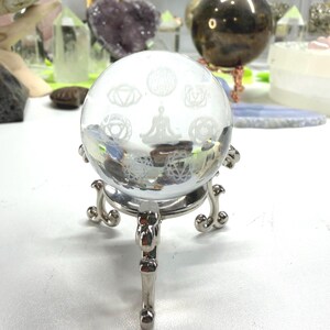 May include: A clear glass sphere featuring etched white symbols of the seven chakras and a central figure in a seated meditation pose. The sphere rests on a polished silver-toned metal tripod stand with curved legs and decorative scroll details.