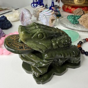May include: A jade green frog statue with a coin in its mouth. The frog is detailed with textured skin and sits on a base. Other decorative items, including crystals and a singing bowl, are visible in the background.