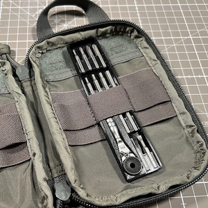 May include: Open olive green tool organizer with a black zipper and handle. Inside, a set of small screwdrivers and a ratchet tool are visible. The label reads "MAXPEDITION AMERICAN DESIGN MADE IN VIETNAM."
