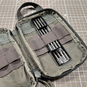 May include: Open, olive green zippered tool case with a black handle. Inside, two black trays hold various silver screwdriver bits. The case has multiple pockets and elastic straps. The text "EXPEDITION AMERICAN DESIGN IN VIETNAM" is visible.