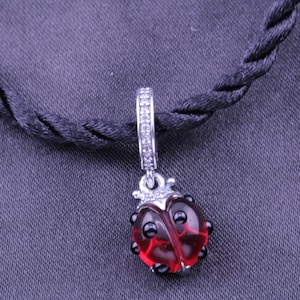 Puede incluir: Un charm de plata en forma de mariquita con alas de vidrio rojo y manchas negras. El charm tiene un bucle de plata en la parte superior y está decorado con pequeños cristales transparentes.