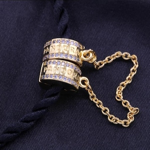 Może przedstawiać: Złoty charms z wygrawerowanym słowem "PANDORA" na dwóch ułożonych na sobie obręczach, ozdobiony małymi niebieskimi kamieniami. Delikatny złoty łańcuszek łączy obręcze. Charms spoczywa na granatowej tkaninie.