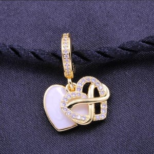 Puede incluir: Pendiente dorado con dos colgantes en forma de corazón. Un corazón es blanco, el otro es un diseño entrelazado incrustado con pequeñas piedras preciosas transparentes. El aro del pendiente también está adornado con piedras preciosas. El fondo es una tela azul oscuro.