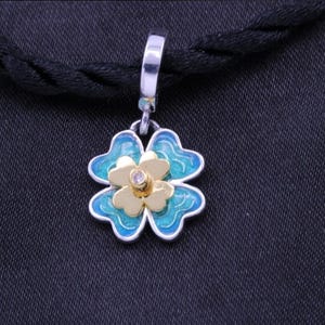May include: A silver-toned clover pendant with a gold center and a small gemstone. The four-leaf clover design features blue enamel accents and hangs from a silver bail. The pendant is displayed against a dark background.