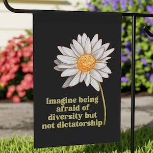 May include: A black garden flag featuring a large white daisy with a yellow center. The flag has the text "Imagine being afraid of diversity but not dictatorship" in gold lettering. The flag is displayed on a black metal stand in a garden.