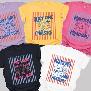 May include: Five short-sleeved crew neck t-shirts in lavender, yellow, pink, black, and white. Each shirt features a central graphic with vertical stripes and text including phrases like Don't Hate the Player Hate the Charleston and Mahjong is Cheaper than Therapy.