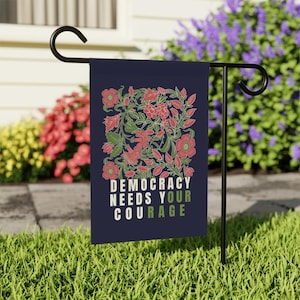 May include: A navy blue garden flag with a floral design in red and green. The flag reads "DEMOCRACY NEEDS YOUR COURAGE" in white and green text. The flag is displayed on a black metal stand in a garden setting.