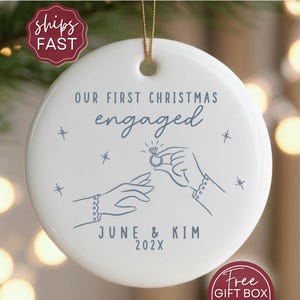 May include: A white ceramic Christmas ornament with the text "OUR FIRST CHRISTMAS engaged" in blue script. The ornament features a line drawing of two hands exchanging a ring, with the names "JUNE & KIM" and the year "202X" below.