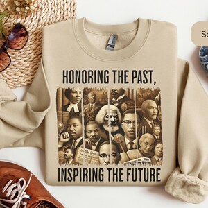 May include: Sand-colored sweatshirt with the text "HONORING THE PAST, INSPIRING THE FUTURE." The graphic features portraits of historical figures. The sweatshirt is paired with blue jeans and brown shoes.
