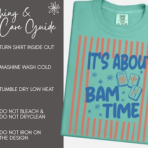 May include: A seafoam green t-shirt featuring a graphic with vertical coral stripes and the blue text IT'S ABOUT BAM TIME, accompanied by mahjong tile illustrations. A gray panel on the left displays a washing and care guide with icons and text.