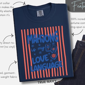 May include: A true navy blue t-shirt featuring a graphic print with vertical coral stripes and the blue text "MAHJONG is my LOVE LANGUAGE" accompanied by three mahjong tile illustrations and small star-like floral accents on the front.