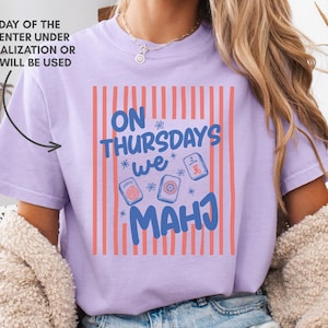 May include: A light purple t-shirt featuring a graphic with vertical coral stripes and the blue text "ON THURSDAYS WE MAHJ" surrounded by small flower icons and three mahjong tiles. The text "ANY DAY OF THE WEEK, ENTER UNDER PERSONALIZATION OR THURS WILL BE USED" is visible.