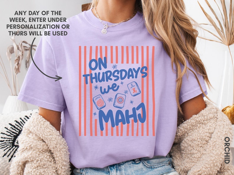 May include: A light purple t-shirt featuring a graphic with vertical coral stripes and the blue text "ON THURSDAYS WE MAHJ" surrounded by small flower icons and three mahjong tiles. The text "ANY DAY OF THE WEEK, ENTER UNDER PERSONALIZATION OR THURS WILL BE USED" is visible.