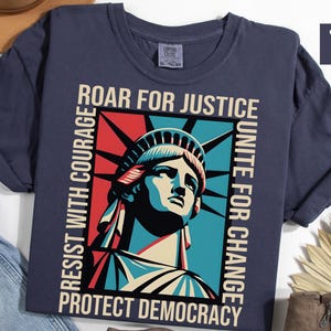 May include: Navy blue t-shirt featuring a graphic of the Statue of Liberty with the words "ROAR FOR JUSTICE", "RESIST WITH COURAGE", "UNITE FOR CHANGE", and "PROTECT DEMOCRACY". The design uses red, blue, and cream colors. The shirt is a comfort colors style.