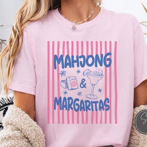 May include: A light pink t-shirt featuring a graphic with vertical pink stripes and the blue text "MAHJONG & MARGARITAS". The design includes illustrations of mahjong tiles, a box, and a margarita glass with a lime wedge and straw.