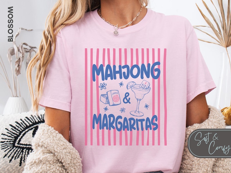 May include: A light pink t-shirt featuring a graphic with vertical pink stripes and the blue text "MAHJONG & MARGARITAS". The design includes illustrations of mahjong tiles, a box, and a margarita glass with a lime wedge and straw.
