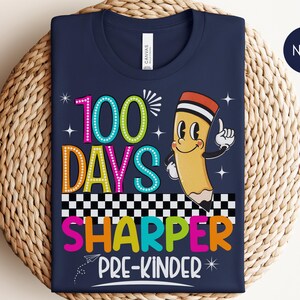 May include: Navy blue t-shirt with a colorful graphic design for pre-kindergarten. The design includes the text "100 Days Sharper Pre-Kinder" with a cartoon pencil illustration. The shirt is folded on a woven basket.