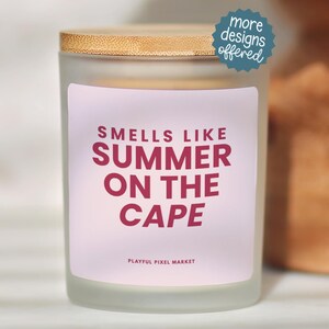 May include: A frosted glass candle jar with a light bamboo lid. The label features the text "SMELLS LIKE SUMMER ON THE CAPE" in bold dark pink letters, with "PLAYFUL PIXEL MARKET" below. A blue circular badge reads "more designs offered".