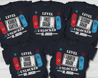 Puede incluir: Camisetas negras con diseños temáticos de juegos para diferentes grados escolares. Cada camiseta presenta un gráfico de controlador, el nivel de grado y el texto "Desbloqueado" con un nombre. Los diseños incluyen "Kindergarten", "Primer Grado", "Segundo Grado", "Tercer Grado" y "Cuarto Grado".