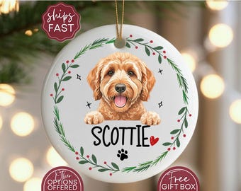 May include: A round, white ceramic ornament featuring a smiling, brown-haired dog with the name "SCOTTIE" below. The ornament is decorated with a green and red floral border, a paw print, and a red heart.