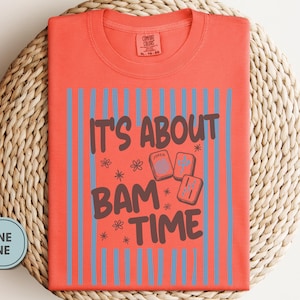 May include: A bright salmon t-shirt featuring a graphic print with vertical light blue stripes and the dark brown text IT'S ABOUT BAM TIME. The design includes small flower icons and three rectangular mahjong tiles with symbols.