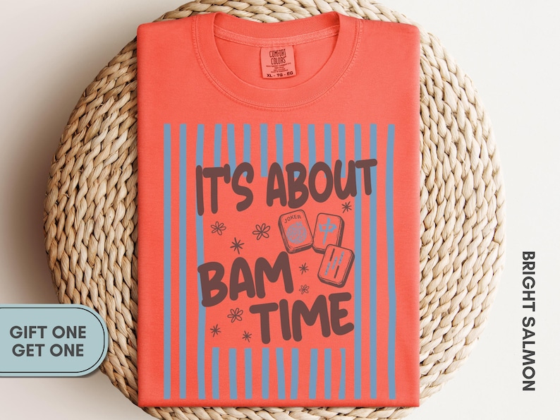 May include: A bright salmon t-shirt featuring a graphic print with vertical light blue stripes and the dark brown text IT'S ABOUT BAM TIME. The design includes small flower icons and three rectangular mahjong tiles with symbols.