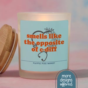 May include: A frosted glass candle with a wooden lid. The candle has a light blue label with the text "smells like the opposite of c-diff" in orange. A stethoscope and floral design are also on the label. The words "Playful Pixel Market" are at the bottom.