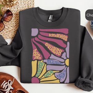 May include: A charcoal gray sweatshirt with a colorful graphic design featuring the words "hope", "aversity", "love", "peace", "inclusion", "equality", and "kindness".
