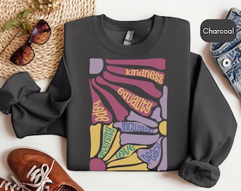 Puede incluir: Una sudadera gris carbón con un diseño gráfico colorido que incluye las palabras "esperanza", "adversidad", "amor", "paz", "inclusión", "igualdad" y "amabilidad".