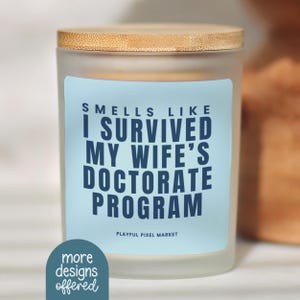 May include: A frosted glass candle with a wooden lid. The candle has a light blue label with the text "Smells Like I Survived My Wife's Doctorate Program." The bottom of the label says "Playful Pixel Market."