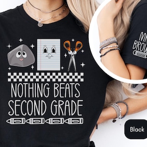 May include: Black t-shirt with a white graphic design featuring a rock, a piece of paper, and scissors. The text "Nothing Beats Second Grade" is below the graphic. The right sleeve of the shirt has the text "Mrs. Brown" in white with a pencil graphic.