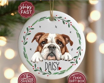 May include: A white ceramic Christmas ornament featuring a brown and white bulldog with the name "DAISY" in black script. The dog is surrounded by a green wreath with red berries and black stars.